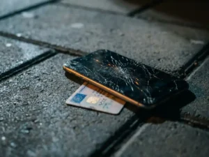 Cracked smartphone screen face-down on concrete, partially covering an EU identity card, dramatic side lighting with amber highlight on card's edge.